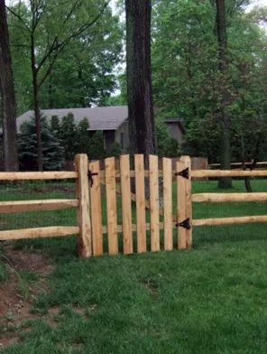 Wooden gate in a split-rail fence in a grassy yard with tall trees and a house in the background