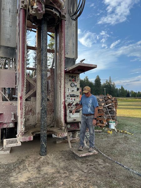 Red drilling rig with stacked pipes, sitting on grass, near a hole in the ground.