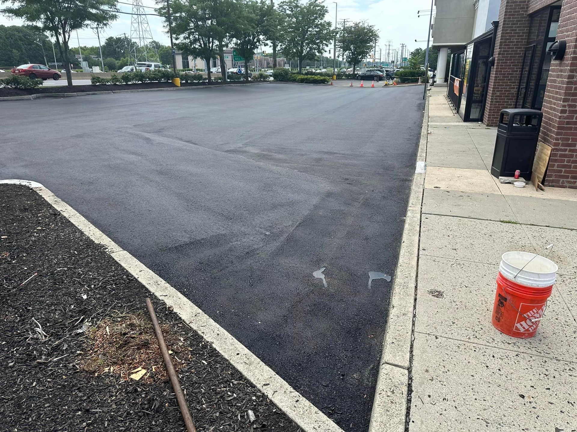 A bucket of paint is sitting on the sidewalk next to a parking lot.