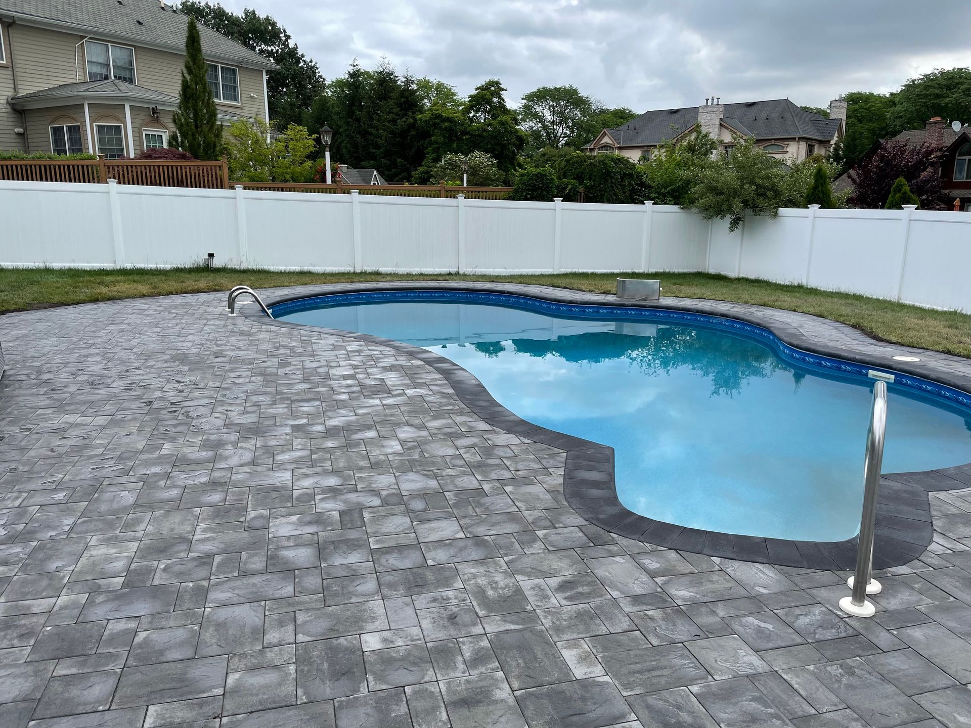 A large swimming pool is surrounded by a brick patio and a white fence.