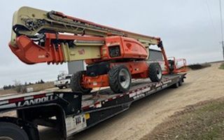 Orange and beige aerial lift on a black flatbed trailer on a dirt road.
