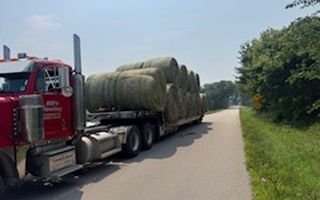 Red semi-truck hauling hay bales on a rural road, sunny day.