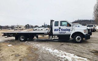 Bill's Towing truck, white and blue, parked on a snowy lot with other vehicles and a trailer in the background.