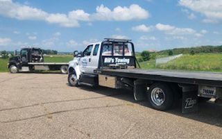 Two flatbed tow trucks parked on pavement under a blue sky, one is white with Bill's Towing on the side.
