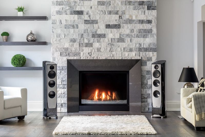 Modern living room with fireplace, speakers, and plush rug. Grey stone wall and wooden floor.
