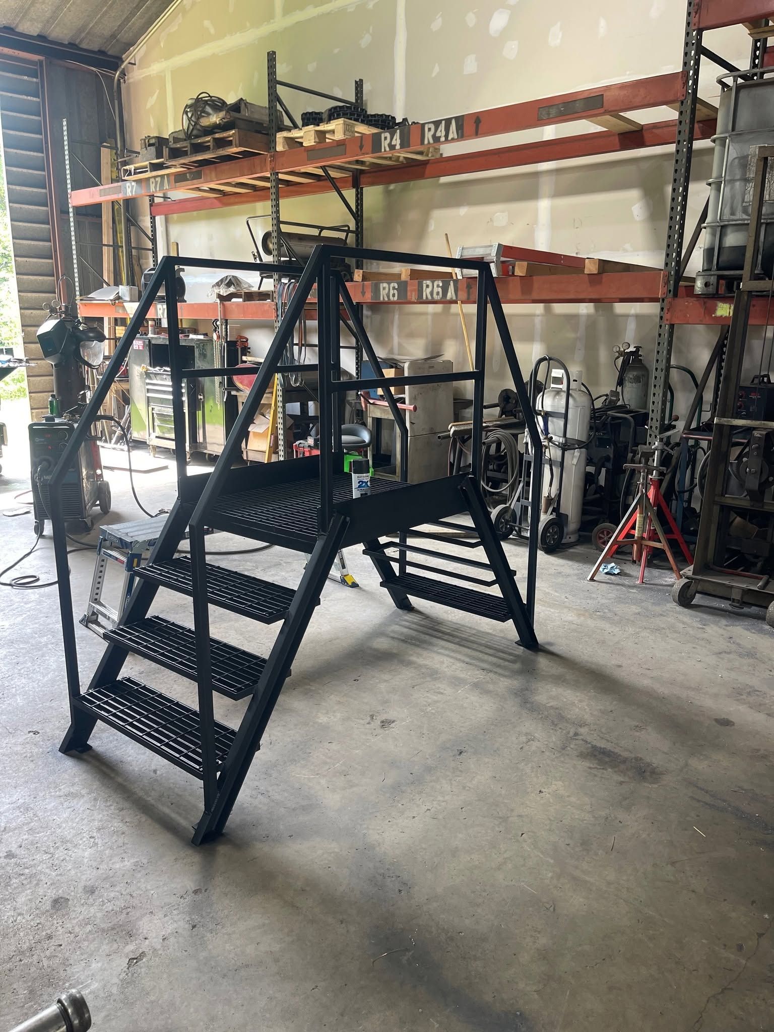 Black metal staircase/platform in a warehouse with shelves and tools in the background.