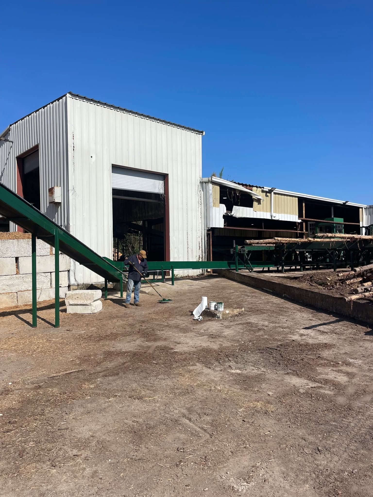 Industrial building with green conveyor pipe and a person standing in a dirt lot under a clear blue sky.