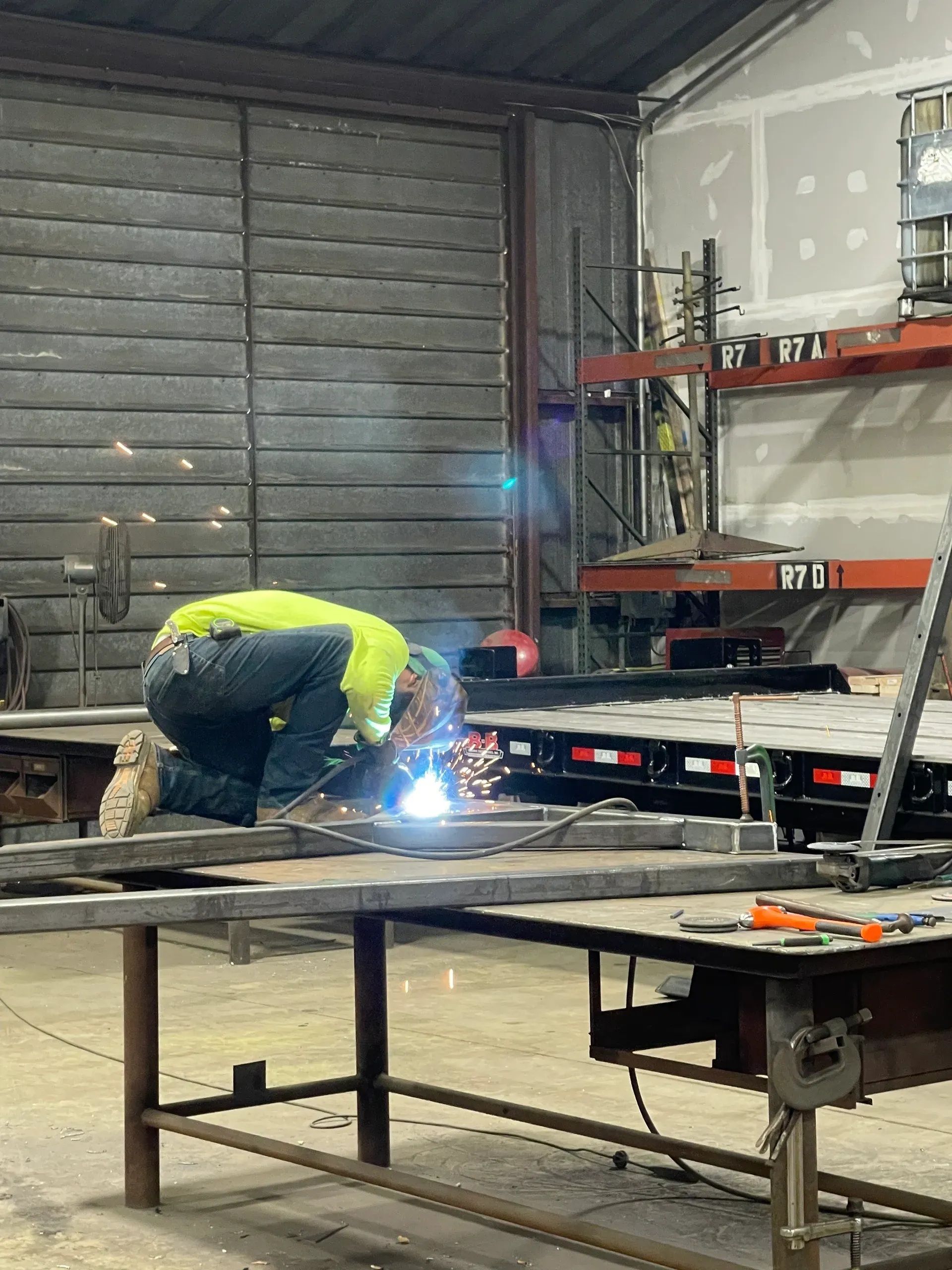Worker welding metal in a workshop, with sparks on a workbench and stacked materials nearby.