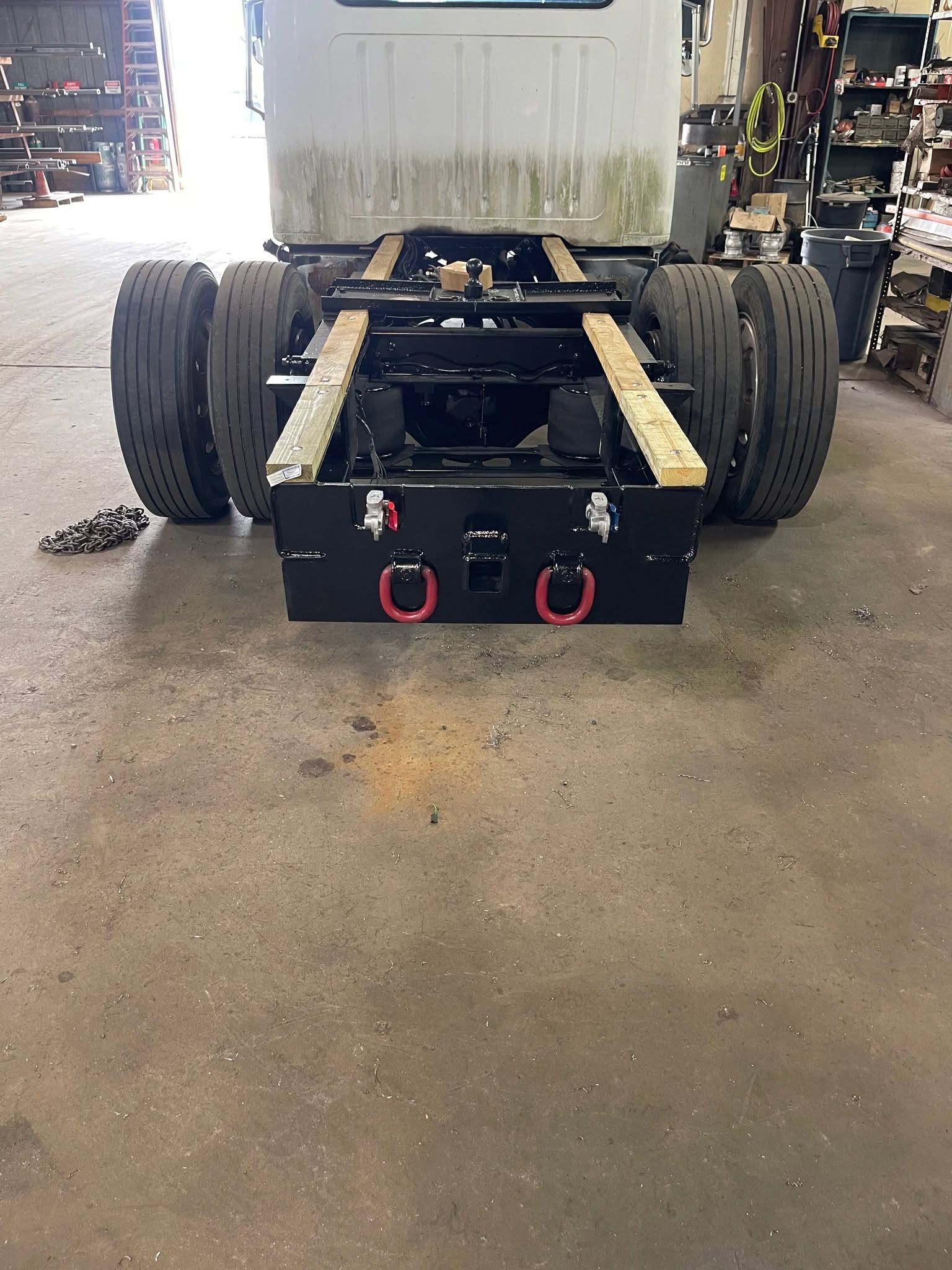 Black truck chassis with large tires and red tow hooks in a garage workshop.