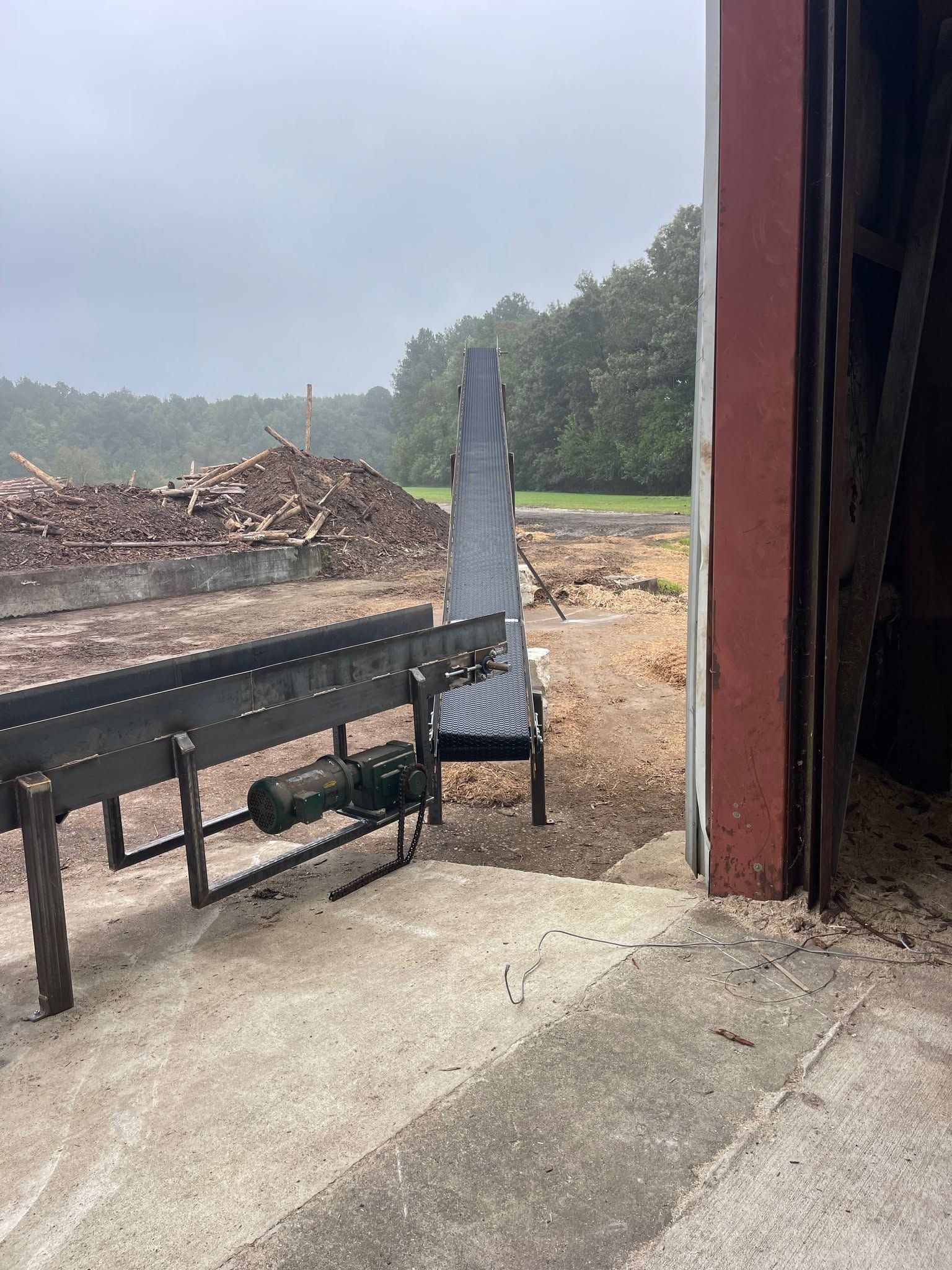 Industrial yard with a conveyor, covered bin, gravel ground, and wood piles under an overcast sky.