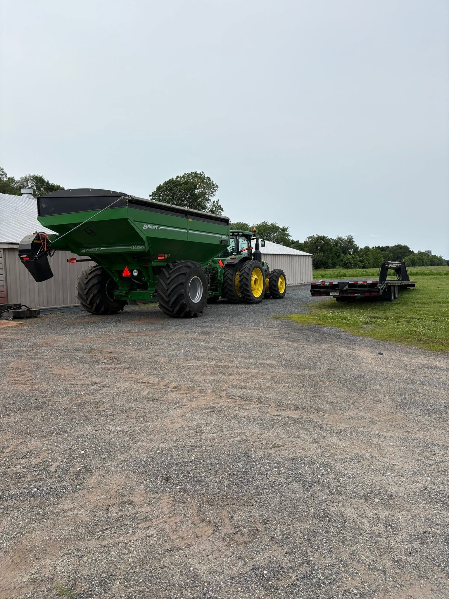 Green grain cart and tractor parked on gravel near a field under overcast sky.
