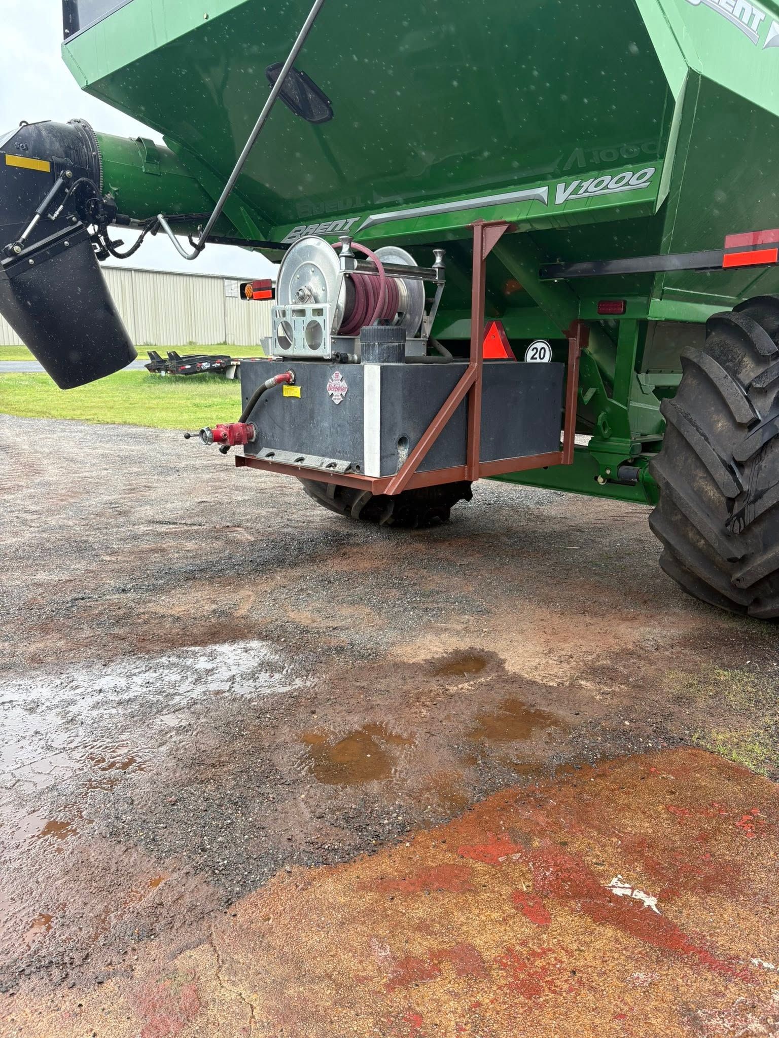 Underneath a green farm machine with black wheel and equipment box on wet gravel.