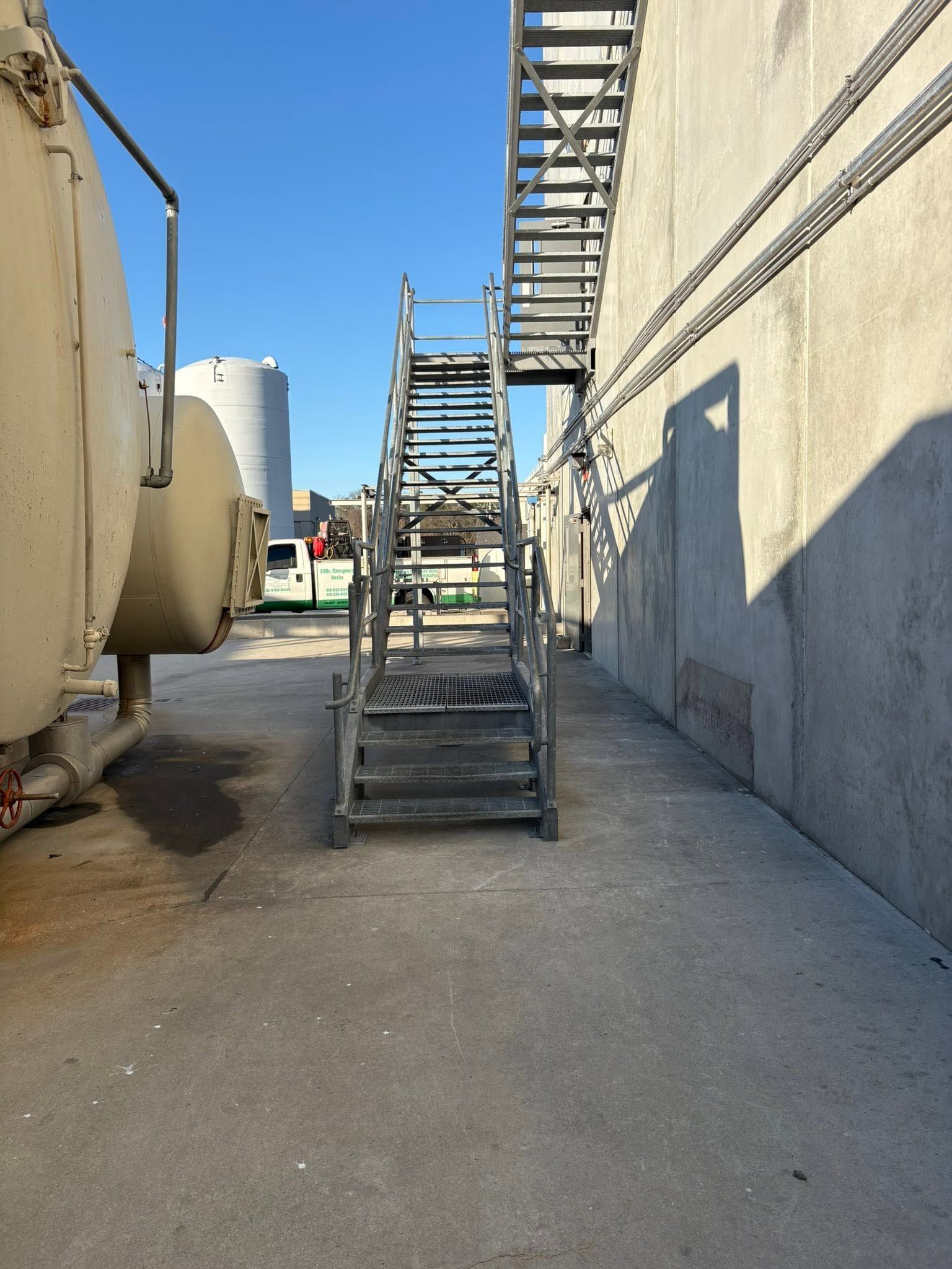 Outdoor industrial alley with metal stairs beside a beige building and large tanks on the left.