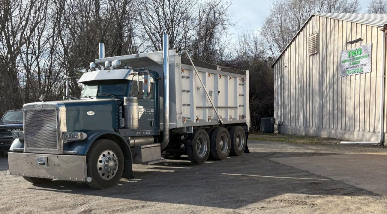 Blue semi-truck with white dump trailer parked beside a barn on a dirt lot.