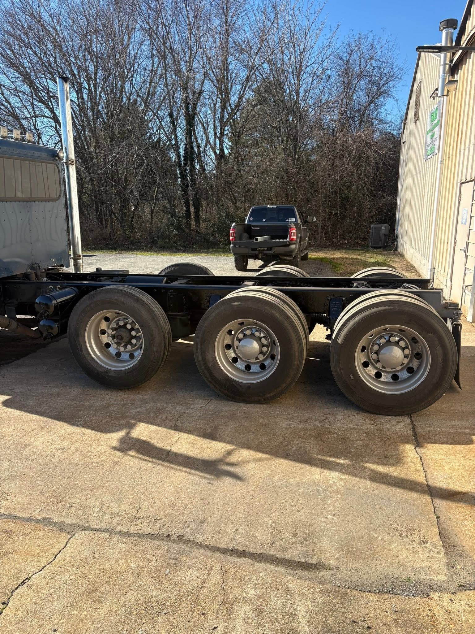 Three-axle trailer chassis parked beside a building on a sunny driveway.