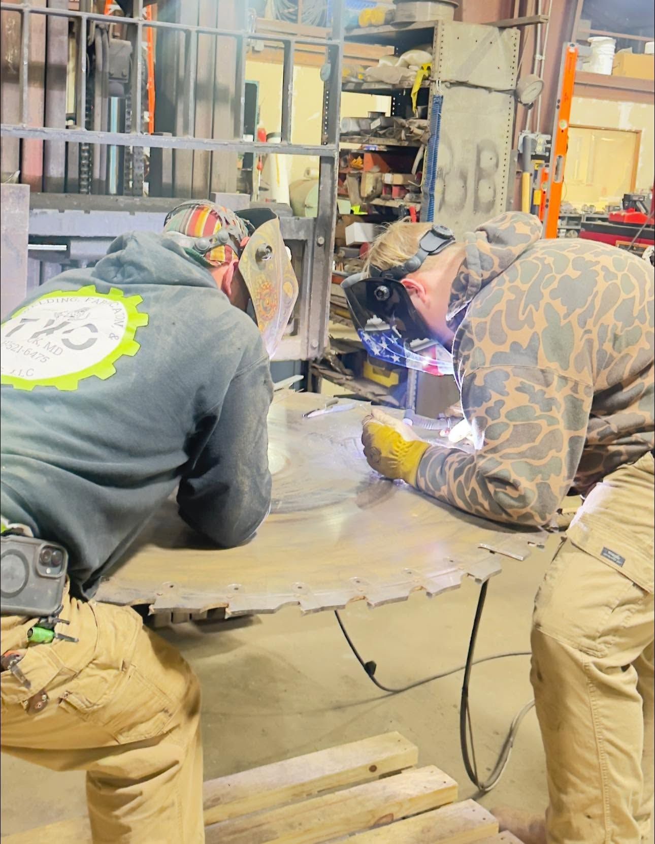 Two workers welding a large metal piece in an industrial workshop, sparks flying.