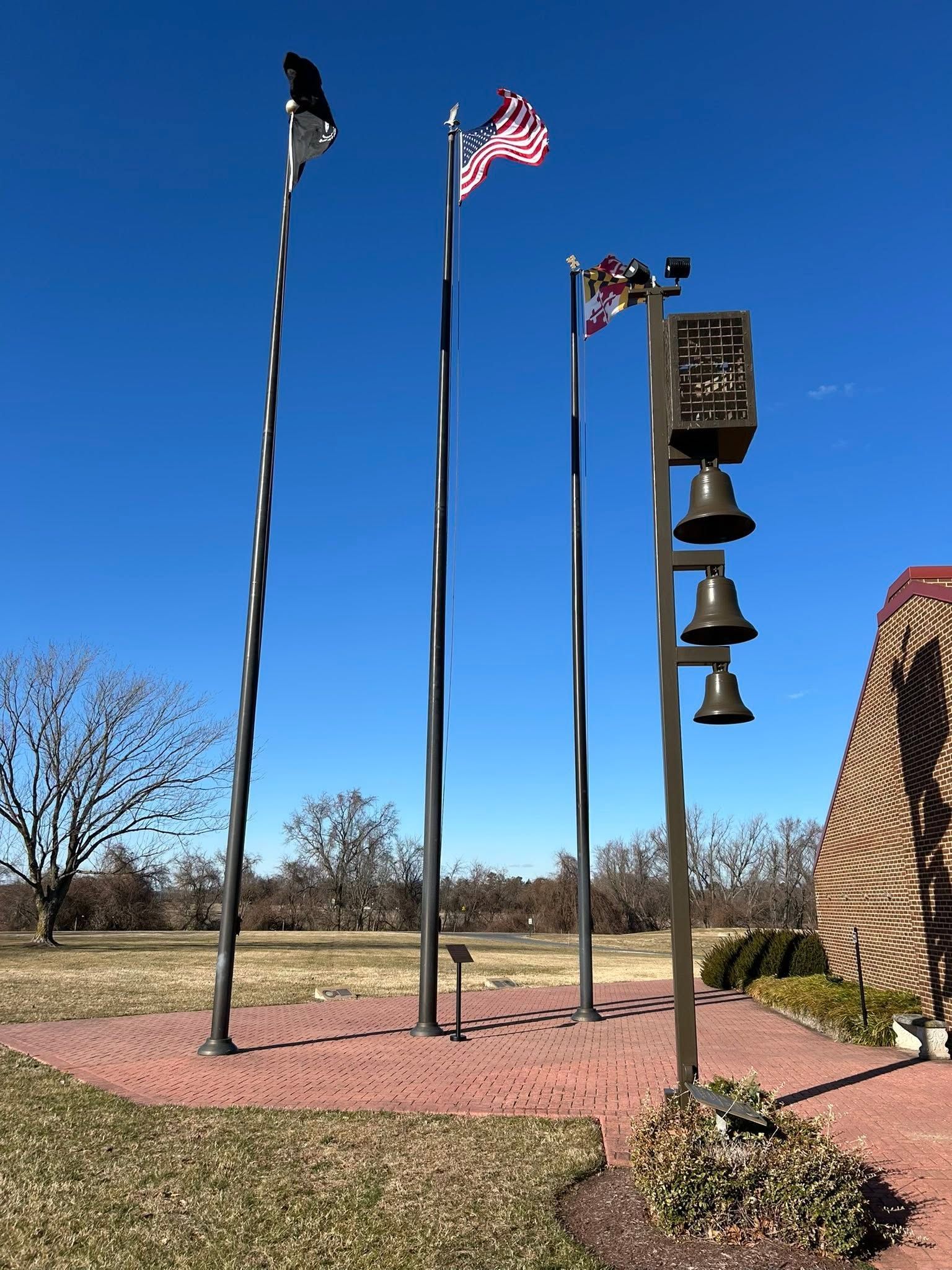 Three tall flagpoles by a bell monument under a clear blue sky in a landscaped outdoor park.