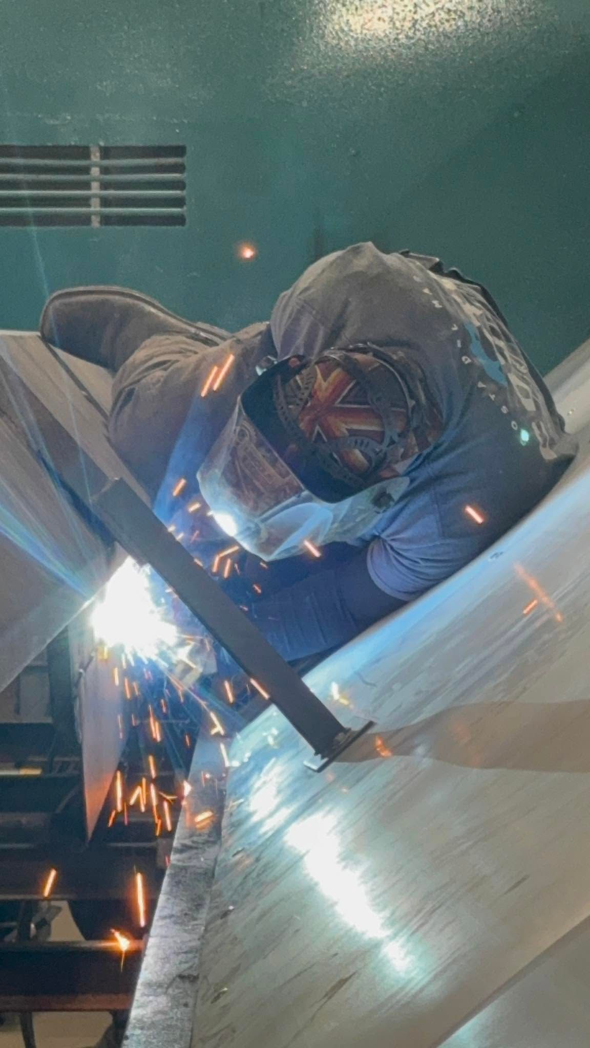 Welder in protective gear sparks metal while working on a large shiny surface indoors.