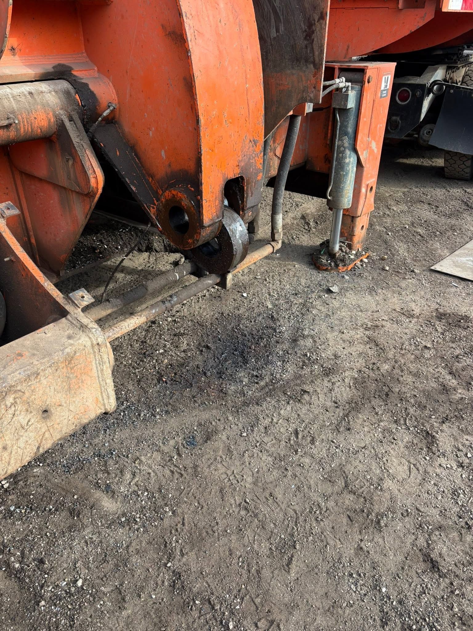 Close-up of an orange snowplow or truck attachment resting on gravel.