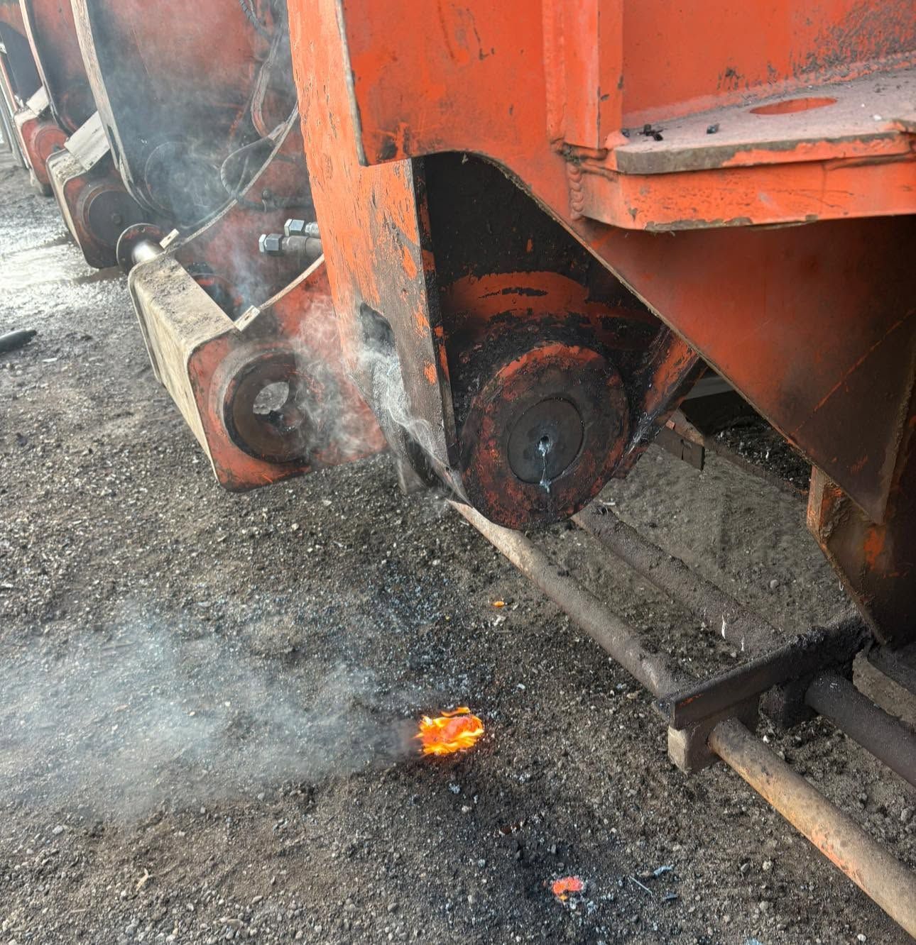 Orange train wheel assembly on gravel, with smoke and a small flame beneath it.