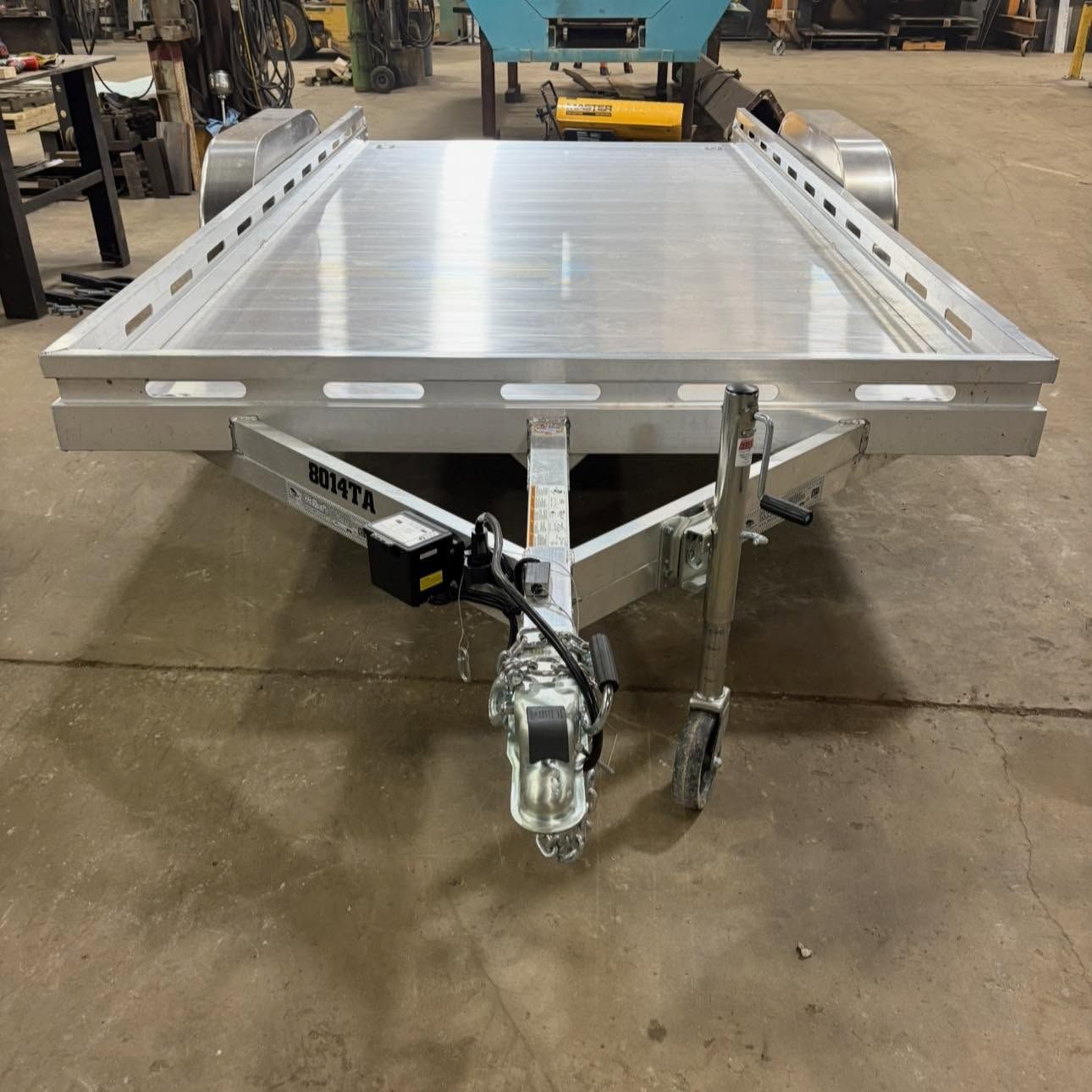 Silver flatbed utility trailer with aluminum deck and hitch in an industrial warehouse.