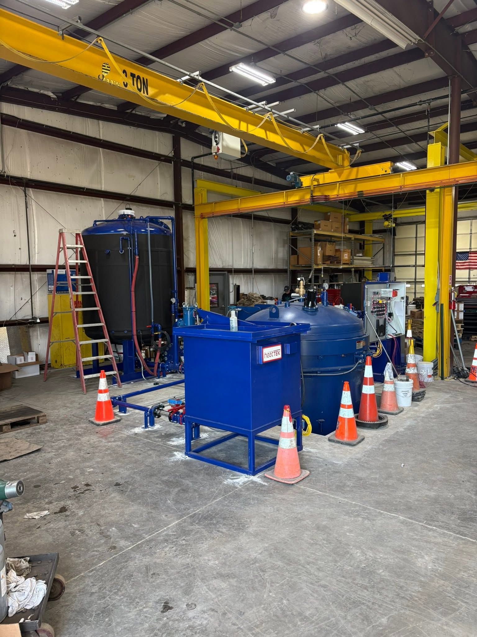 Industrial warehouse with blue tanks, safety cones, and overhead yellow crane equipment under installation.