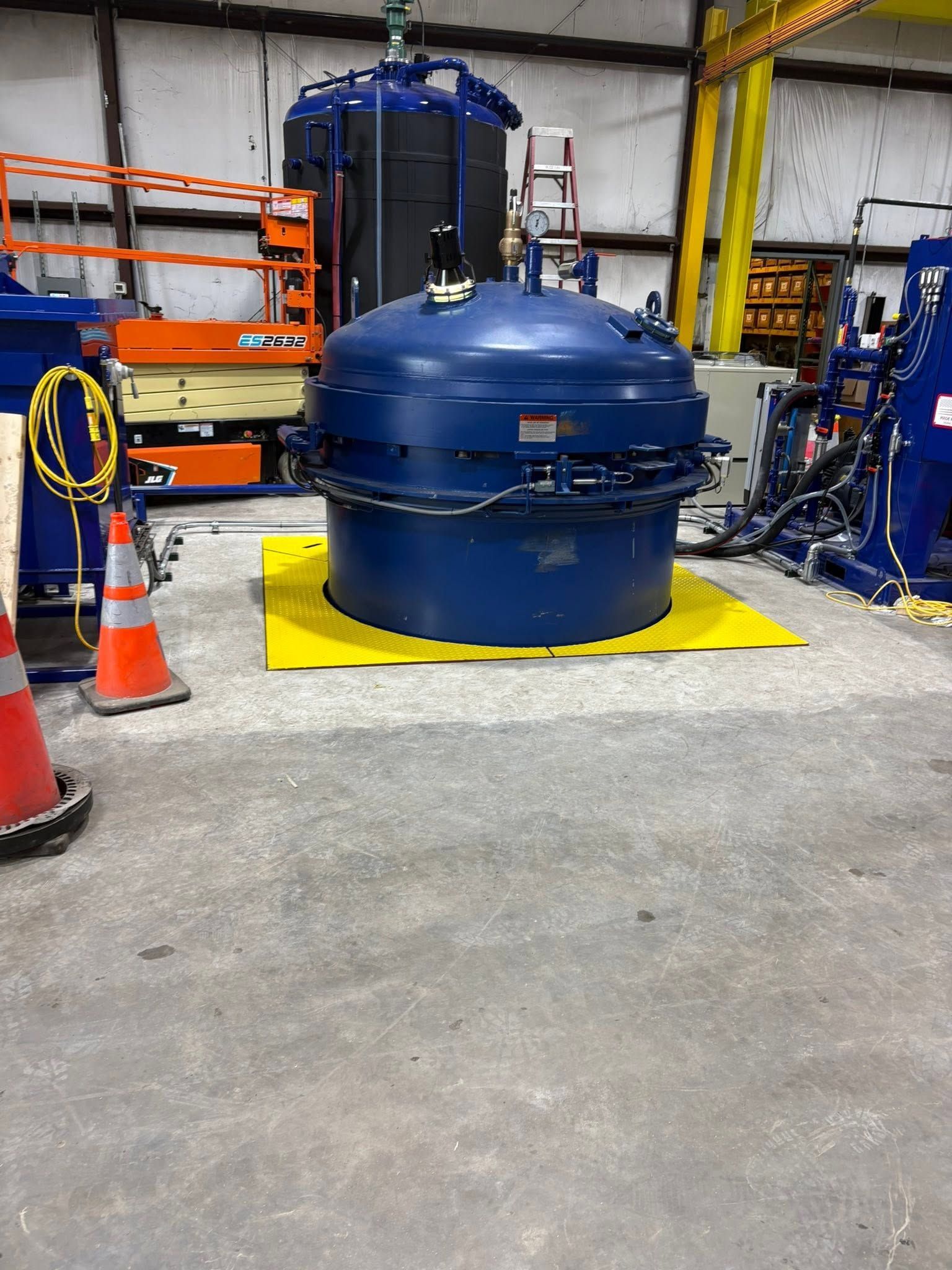 Blue industrial machine in a warehouse, surrounded by orange cones and storage racks.