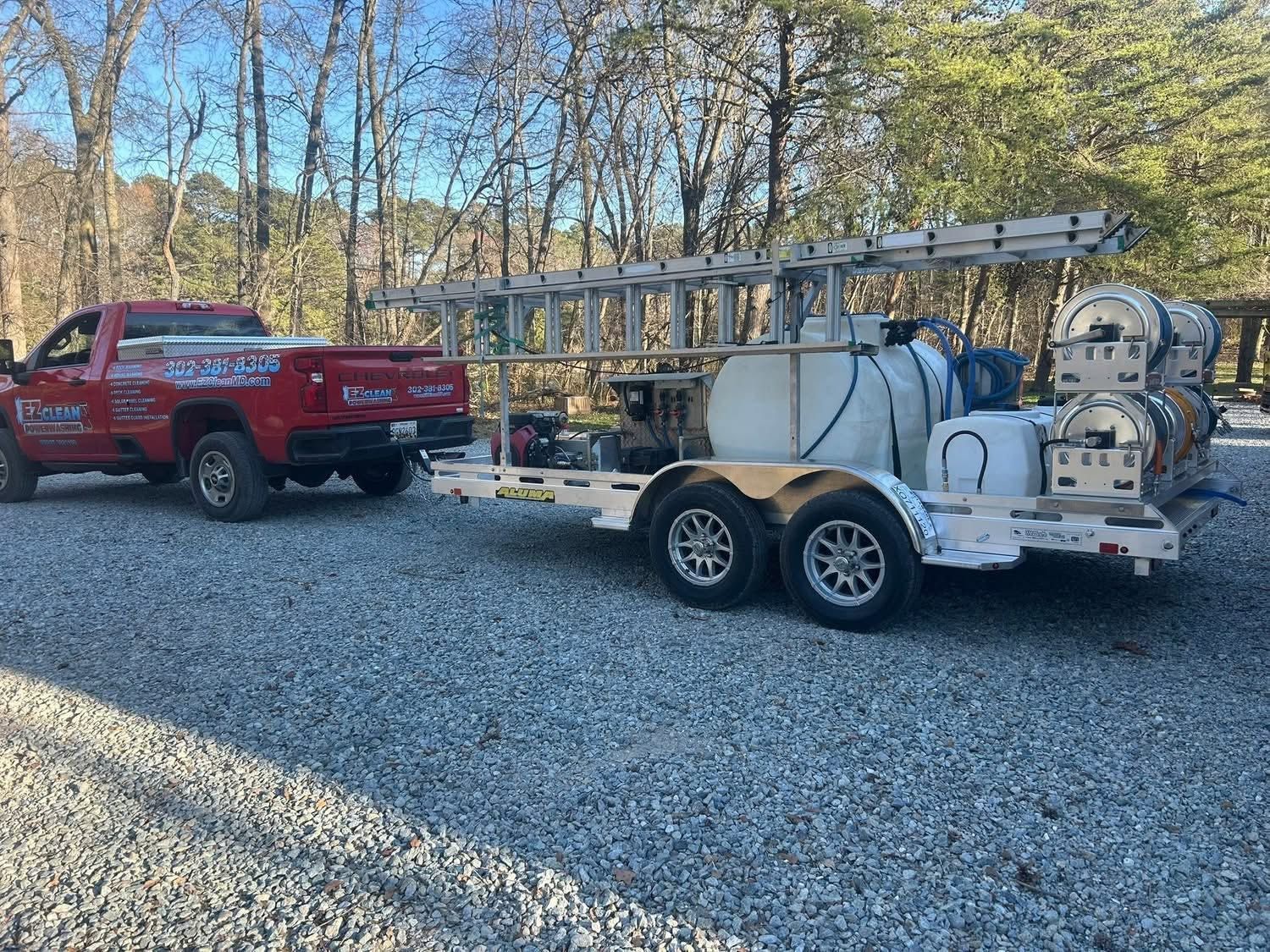 Red pickup towing a white utility trailer with tanks and ladder in a gravel lot beside trees.