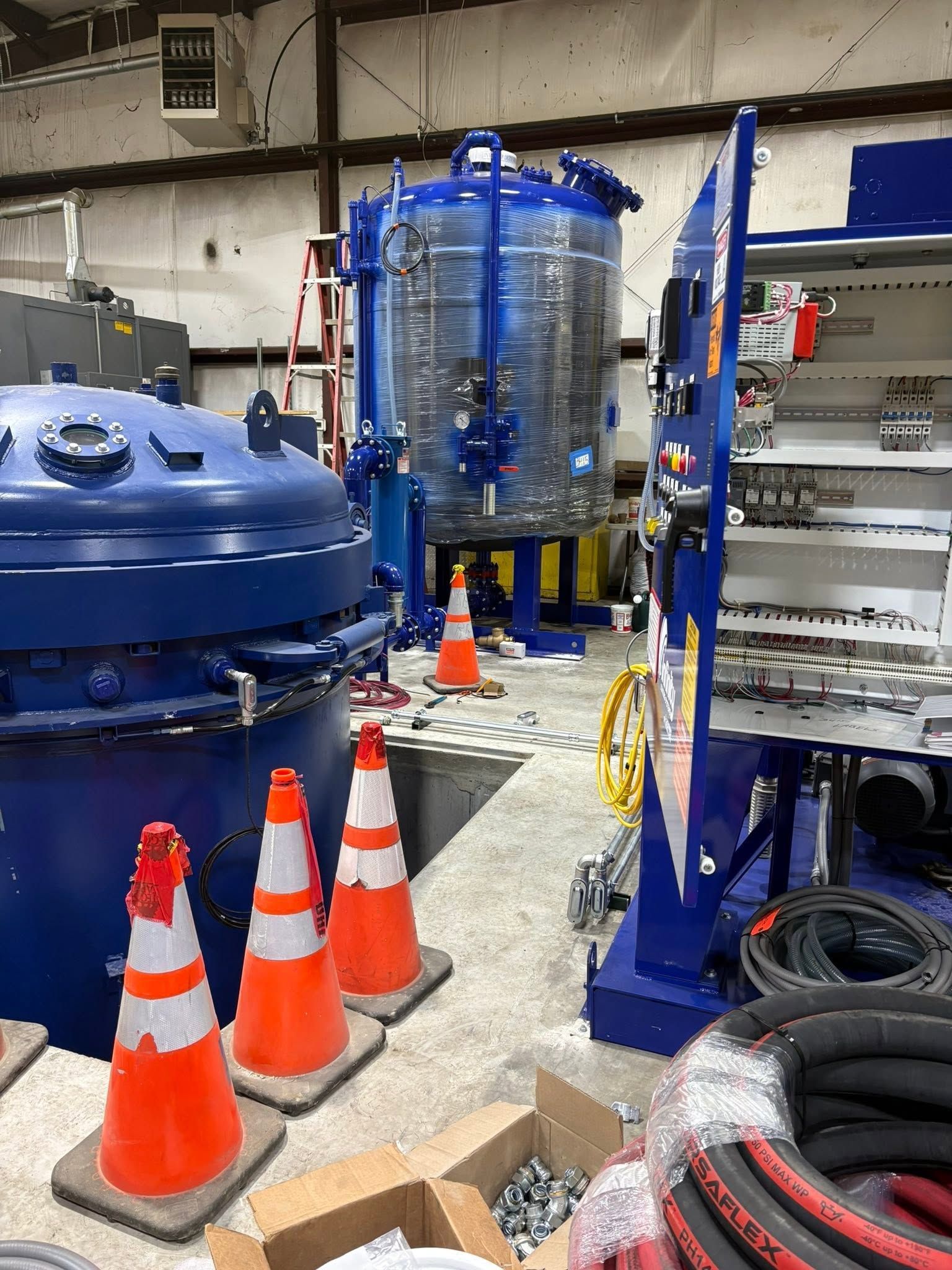 Industrial facility with blue machinery, clear tank, orange cones, and coiled hoses on the floor.