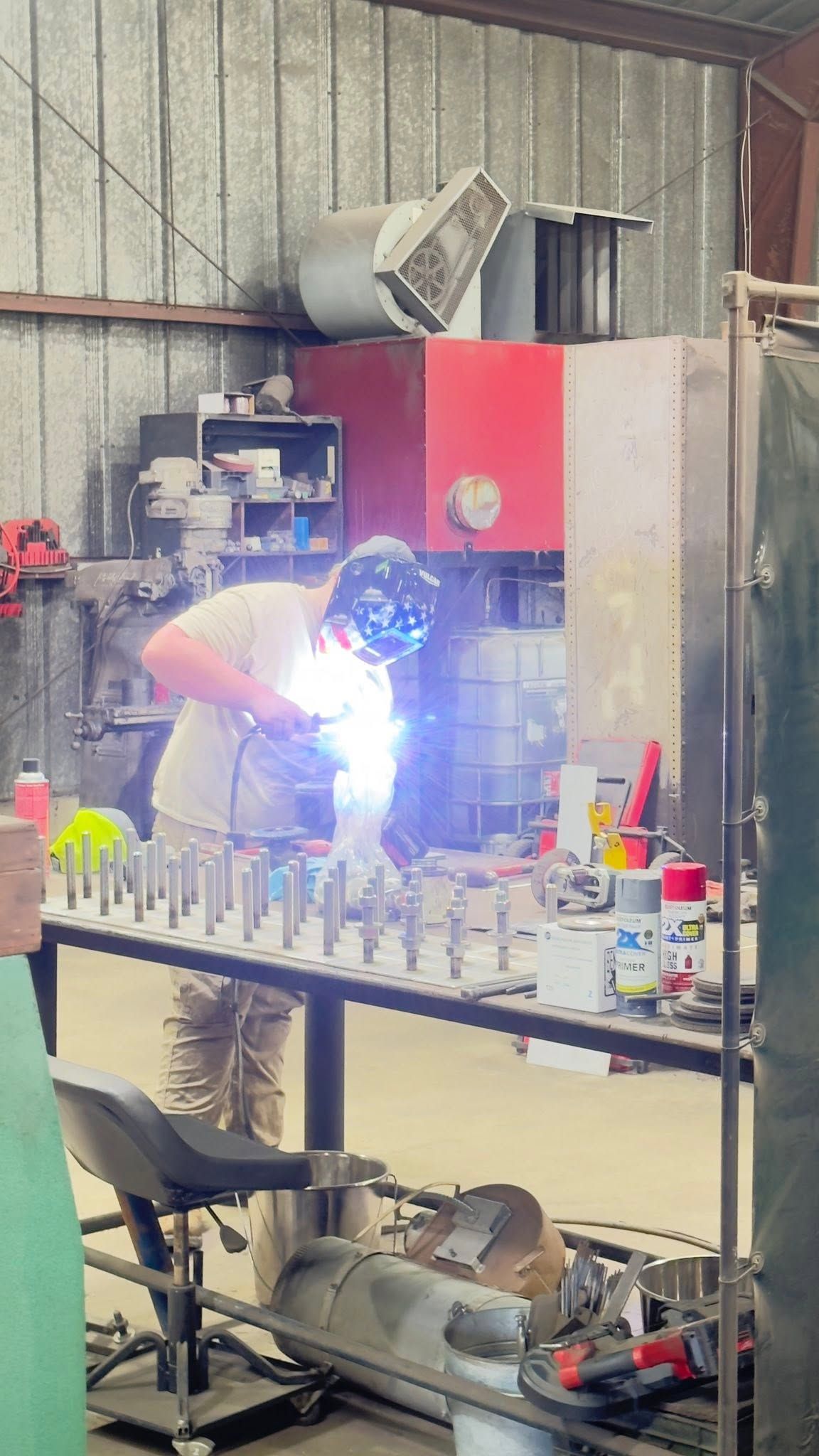 Person welding at a workbench in a metal shop, bright sparks under a red cabinet.
