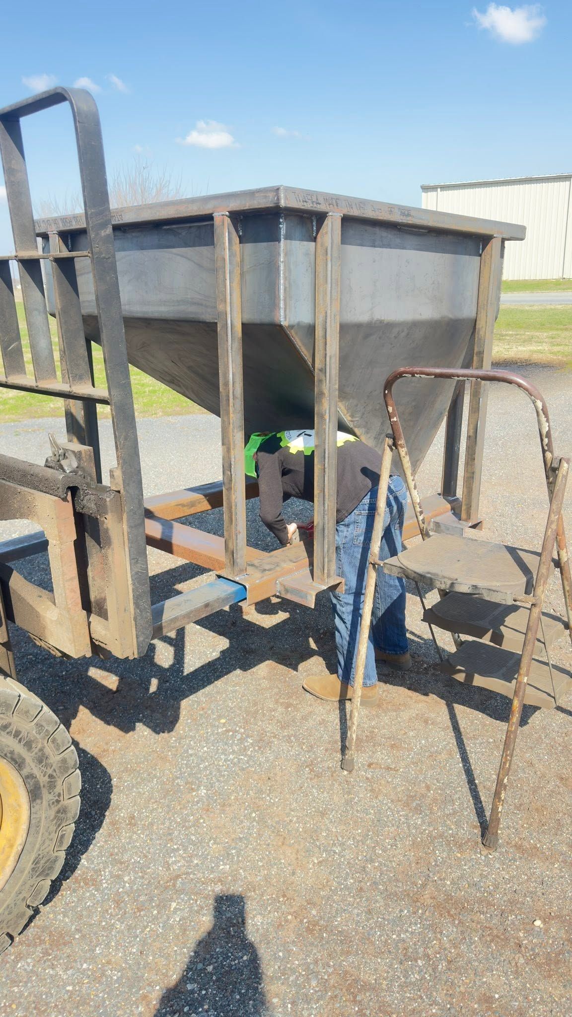 Metal farm hopper on a trailer with blue piping and a yellow wheel, parked on gravel outdoors.