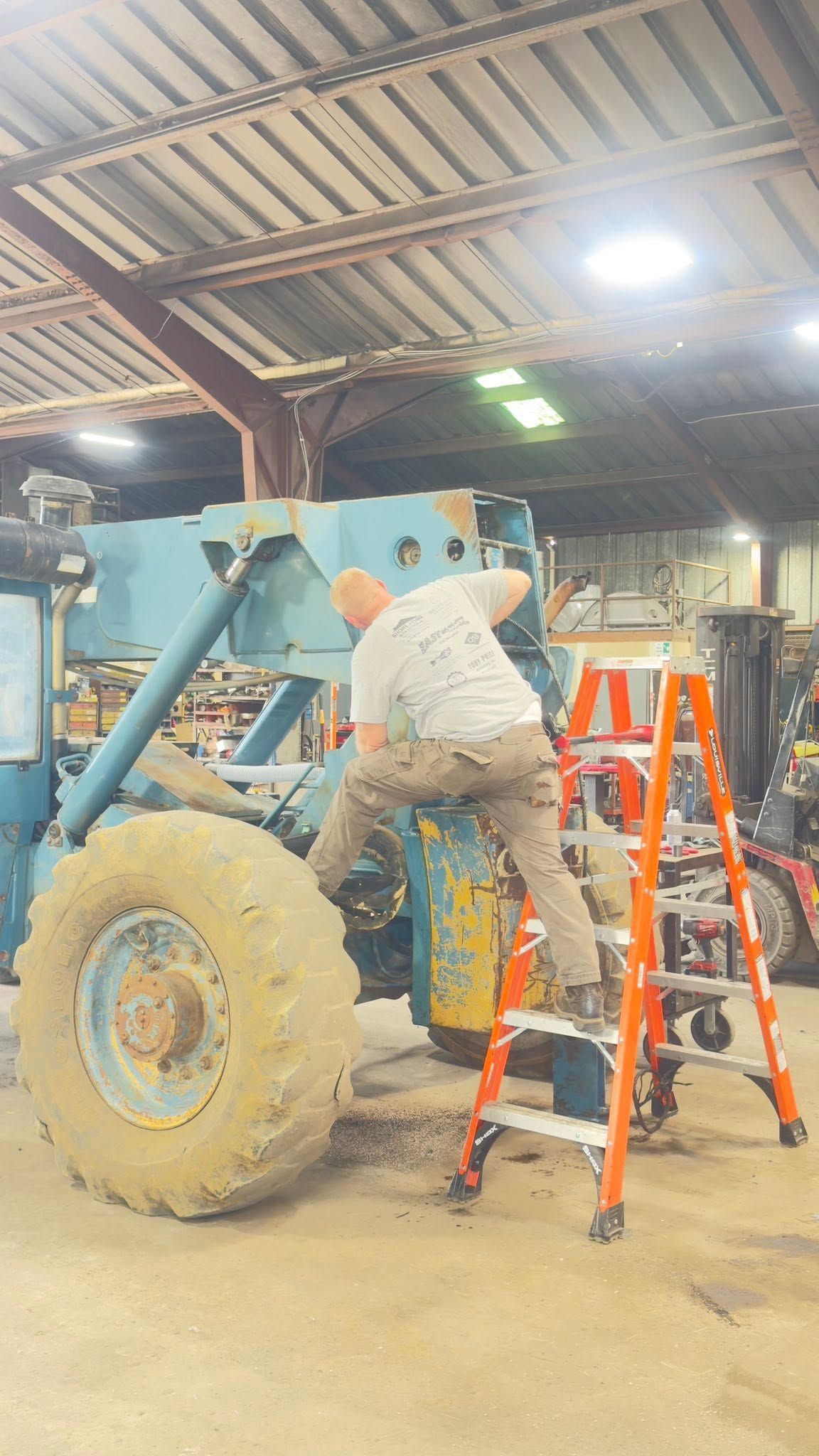 Mechanic working on a blue tractor beside orange ladders inside a repair shop.