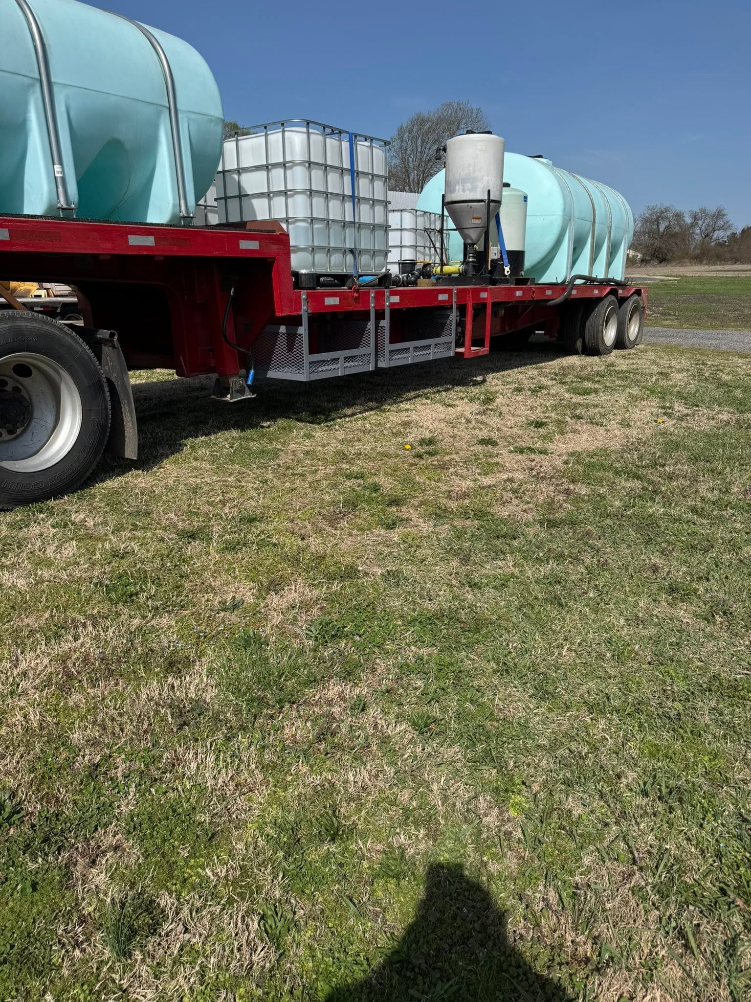 Red flatbed truck carrying large blue tanks and white containers in a grassy field under a clear sky.
