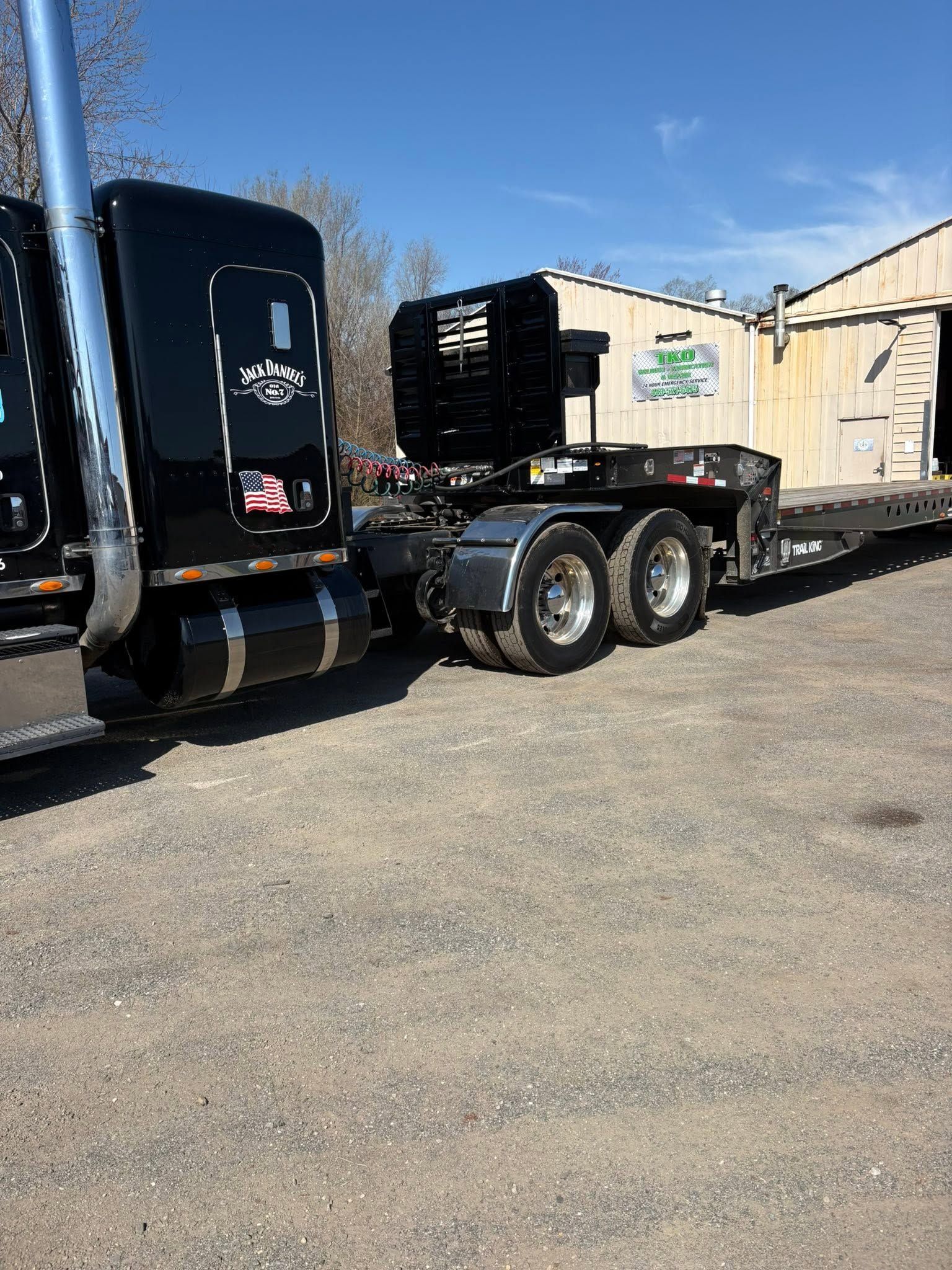 Black semi-truck cab with flatbed trailer parked beside a beige building on a sunny day.