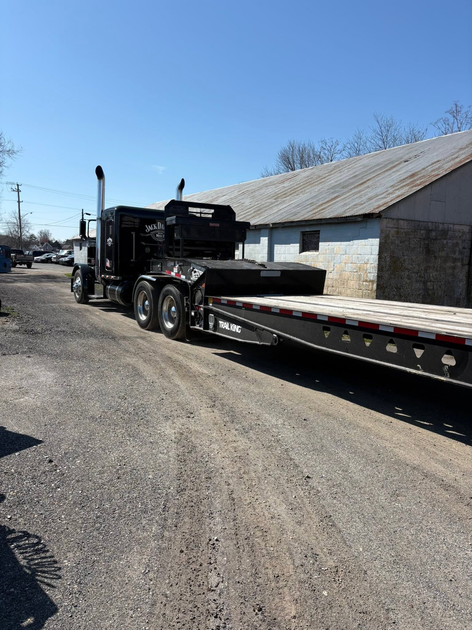 Black flatbed tow truck parked on a gravel lot beside a metal building under a clear blue sky.