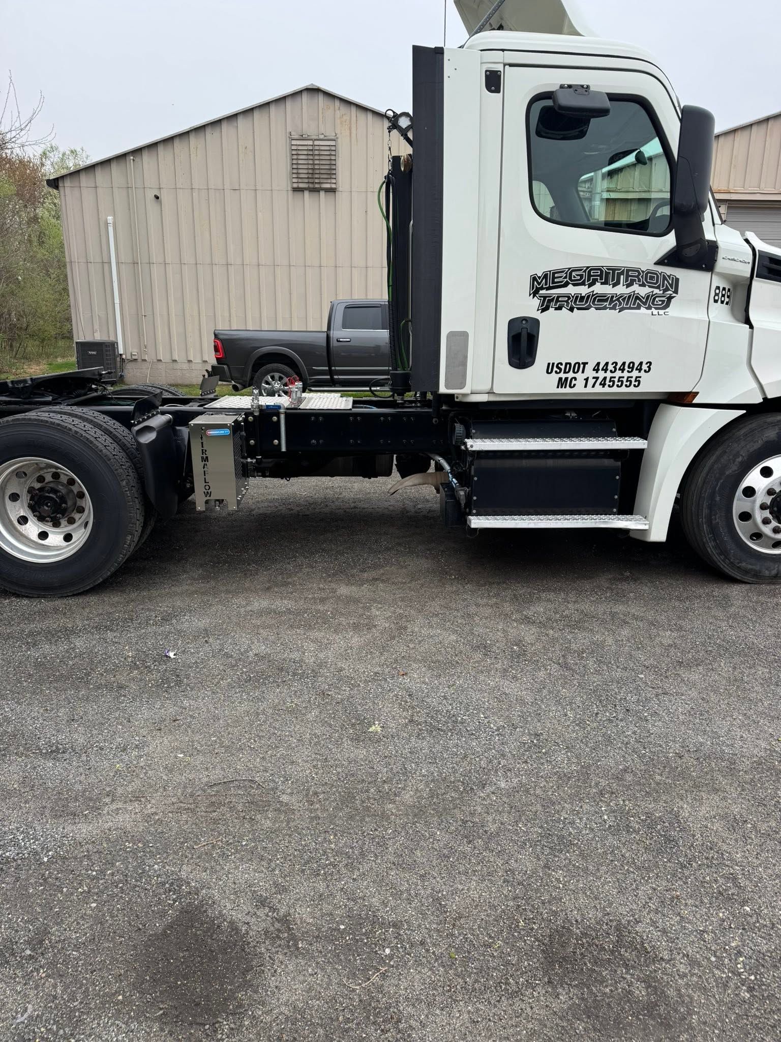 White semi-truck cab parked on gravel beside a beige shed.