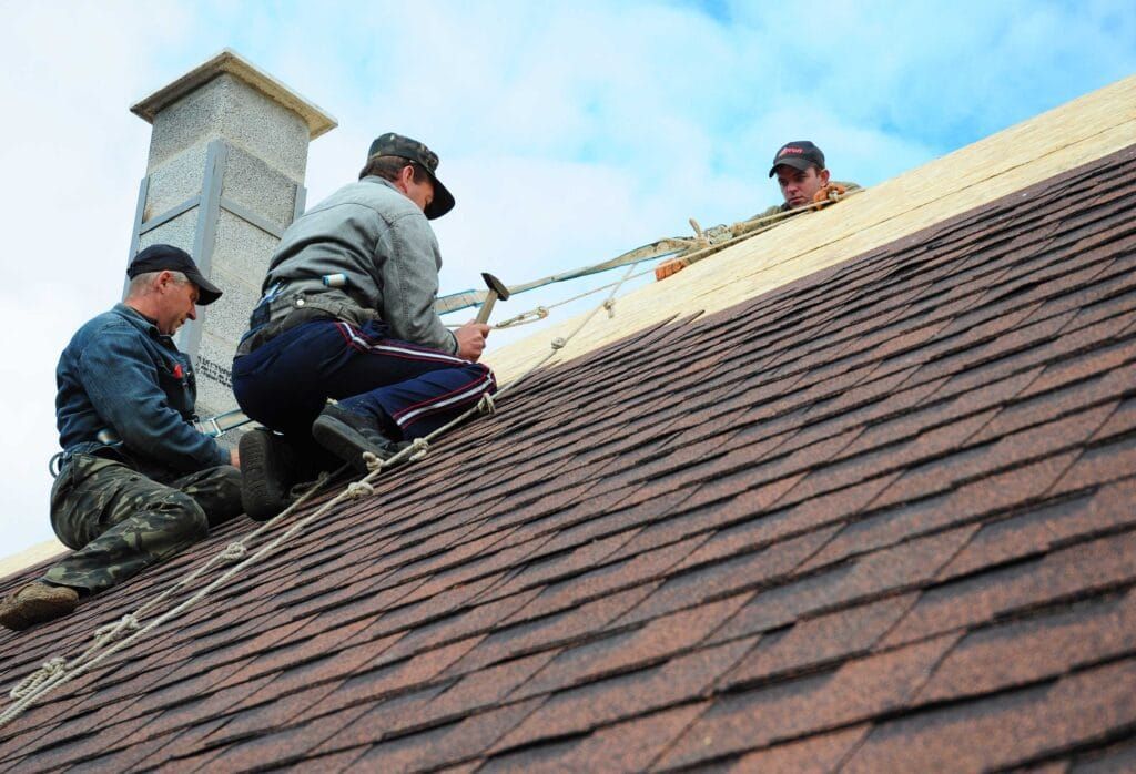 Workers installing shingles on a steep roof under a blue sky