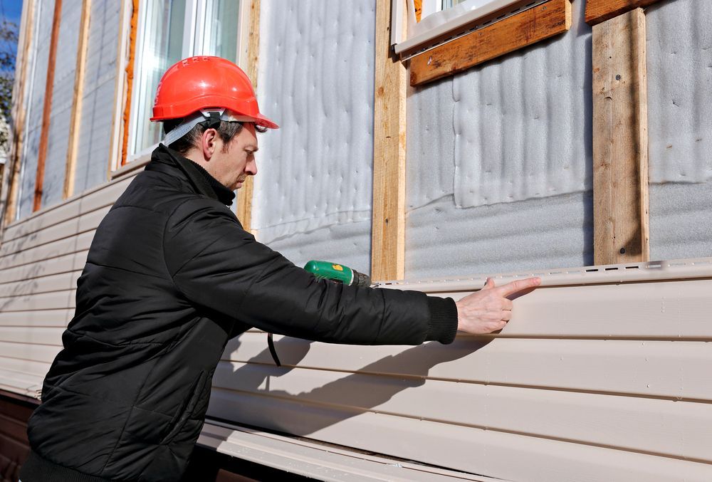Worker in red hard hat installs beige siding on a building exterior.