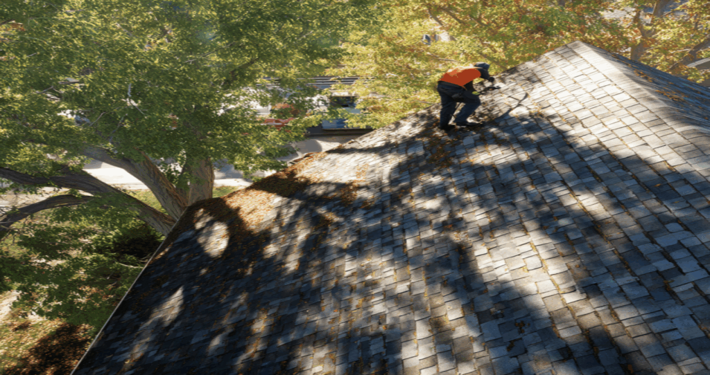 Worker in an orange shirt repairing a sunlit shingled roof beside trees