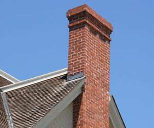 Red brick chimney on a house with a gray shingled roof against a blue sky