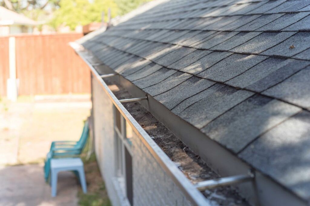 Close-up of a shingled roof with a gutter along the edge, beside a backyard fence and chair.