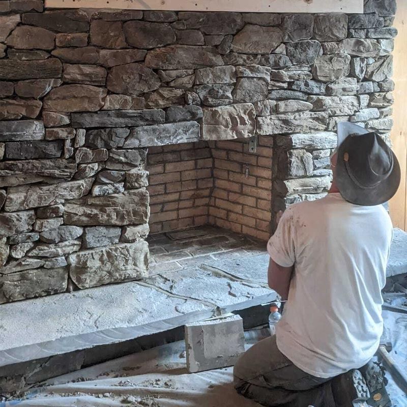 Person kneeling before a stone fireplace under construction, with stacked stone and brick firebox.