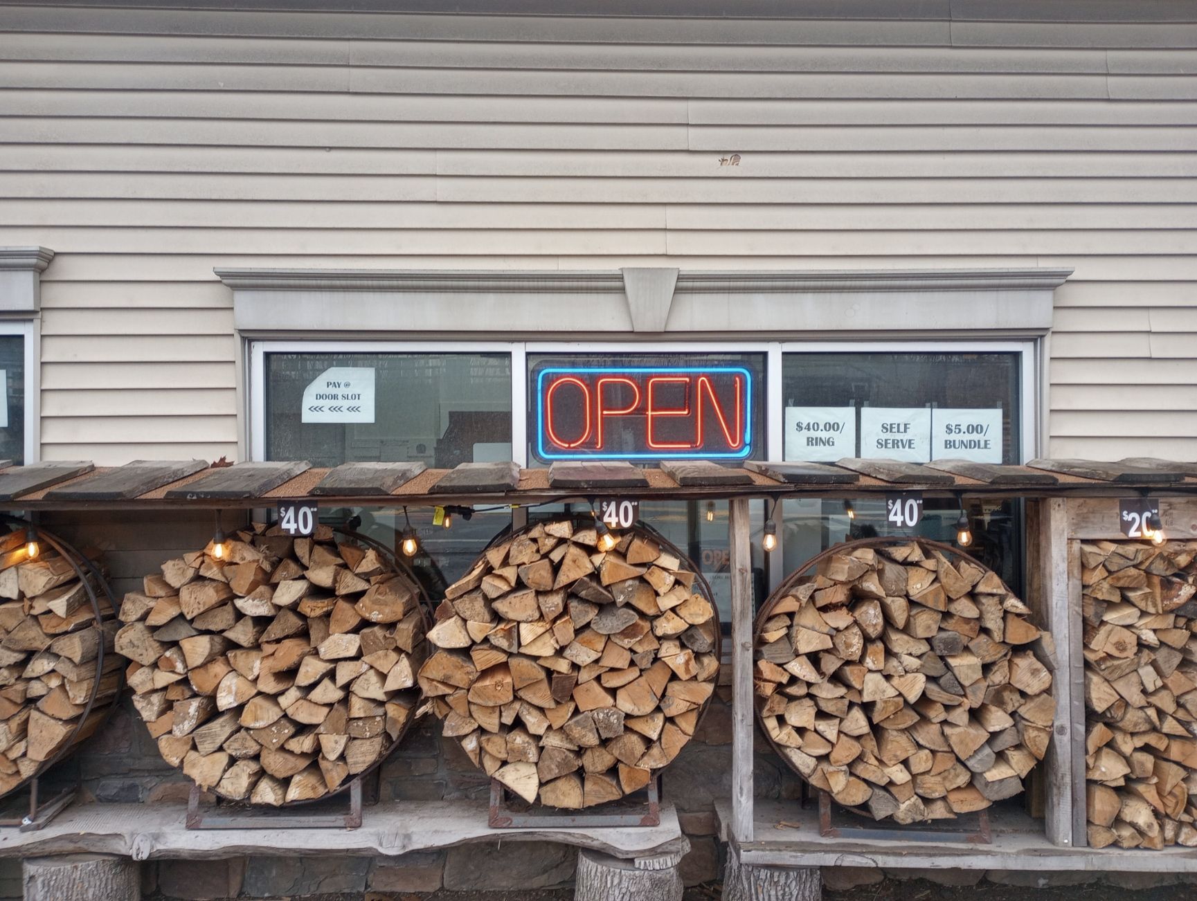 Firewood stacked in racks outside a shop with an illuminated OPEN sign above the window.