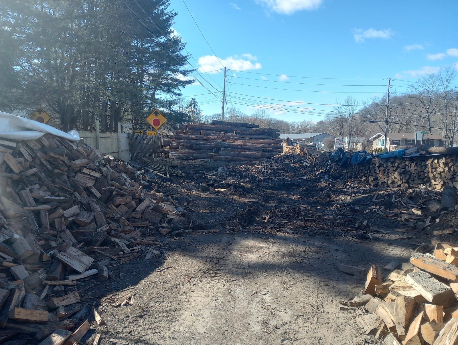 Large piles of chopped wood line a gravel path under a bright blue sky near a roadside sign.