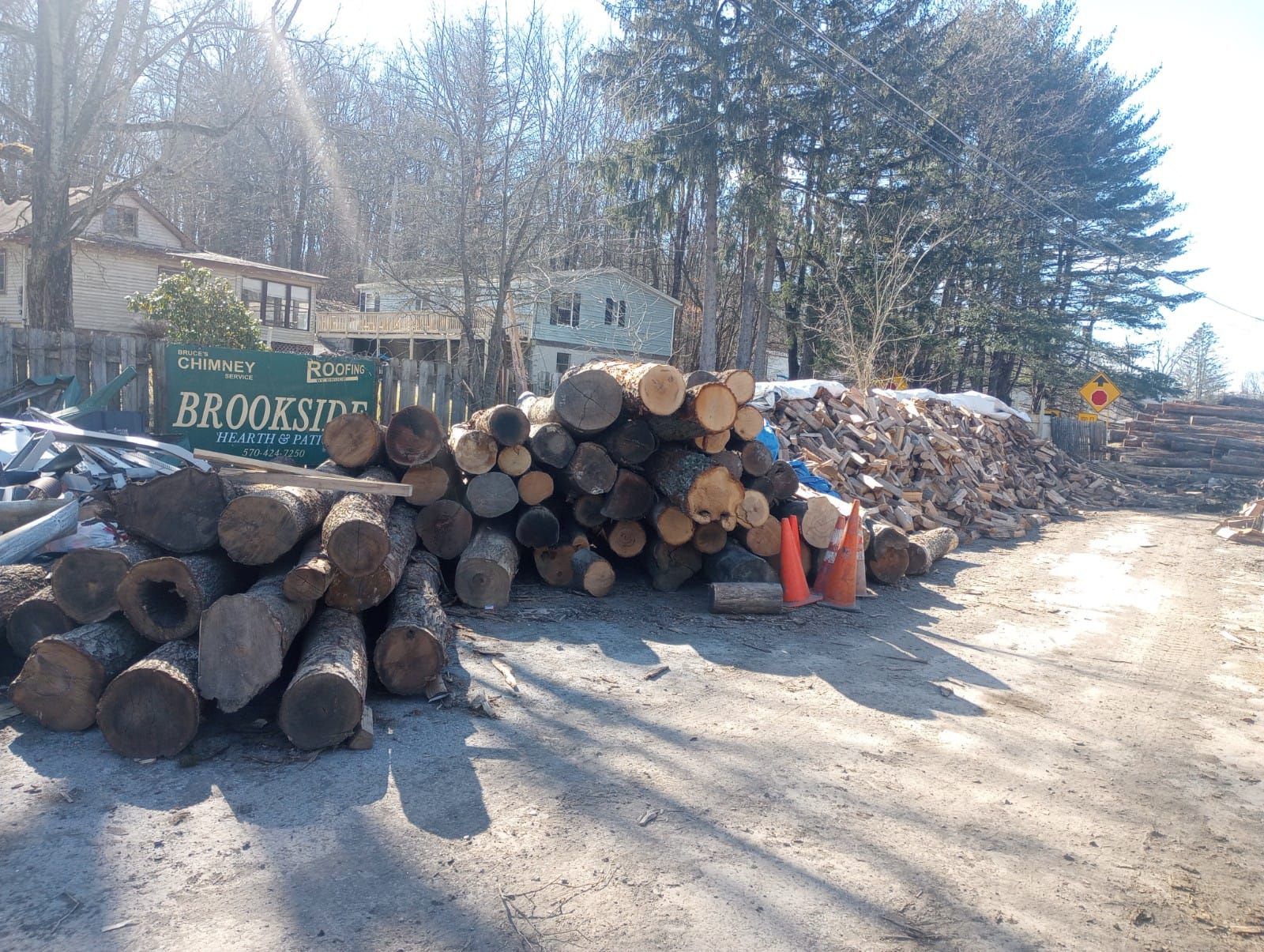 Stacked cut logs beside a dirt road, with trees and a Brooks sign in the background