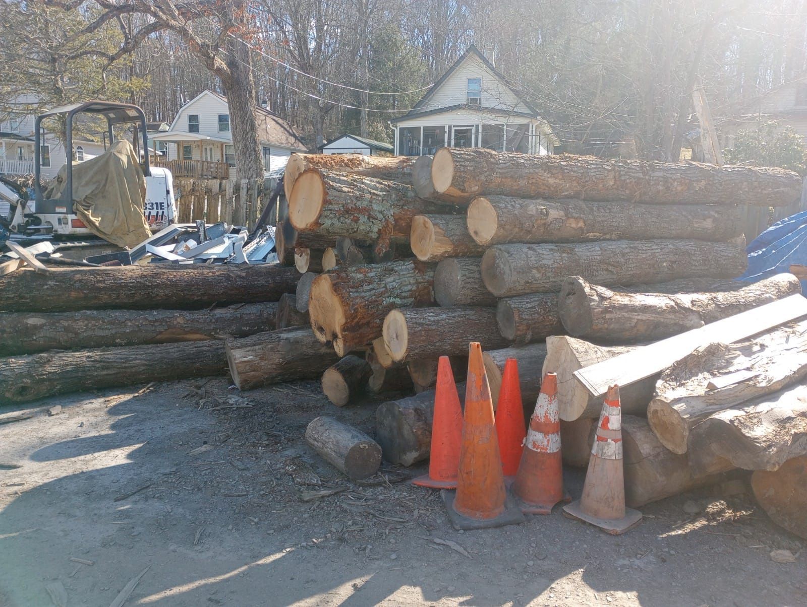 Stacks of cut logs and orange traffic cones along a roadside in a wooded residential area