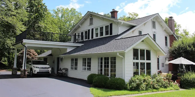 White house with black roof, carport, and lush green trees in a sunny residential setting