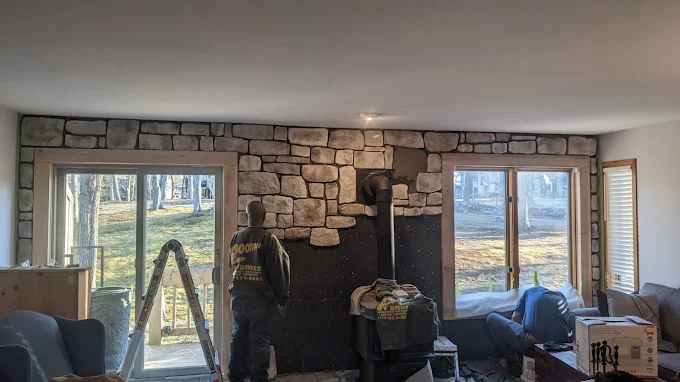 Stone fireplace in a cozy room with large windows and a ladder leaning near the left window.