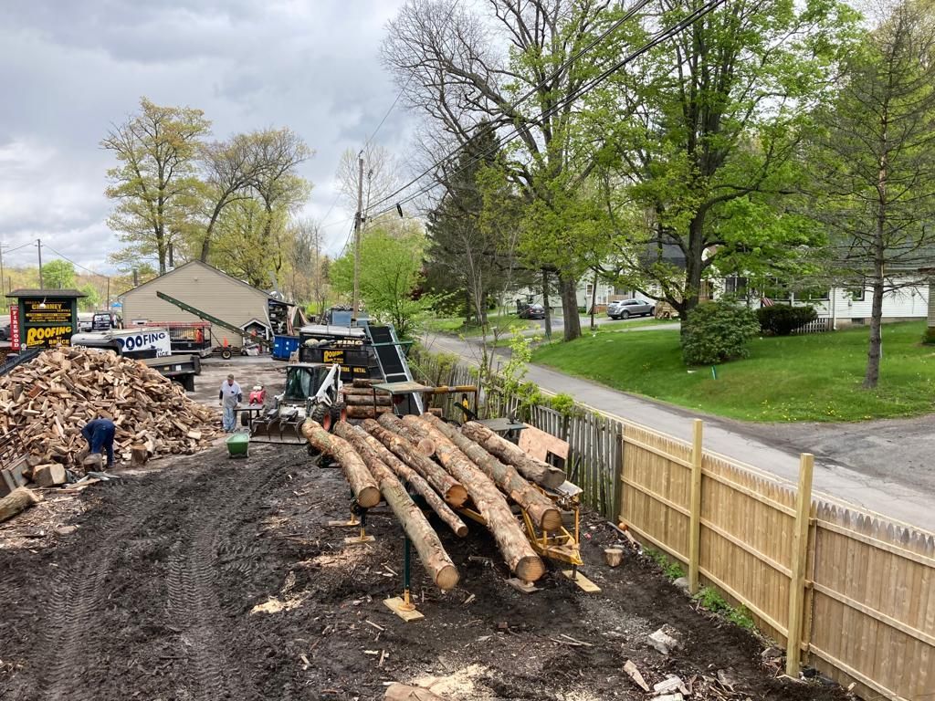 Construction site with stacked logs, dirt piles, and machinery beside a fenced road in a leafy neighborhood