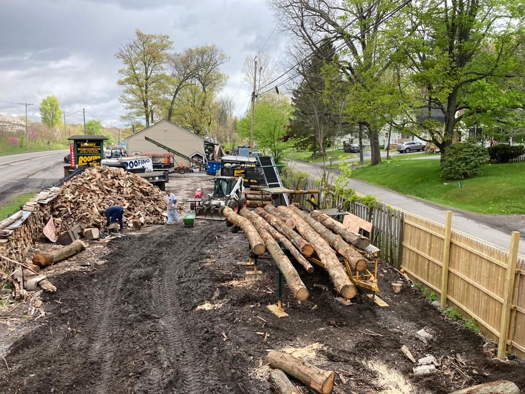 Roadwork site with piles of cut logs, dirt, and construction vehicles beside a fenced grassy area